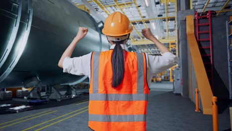 vista trasera de una ingeniera con casco de seguridad levantando las manos celebrando mientras trabajaba en una fábrica de fabricación de tubos