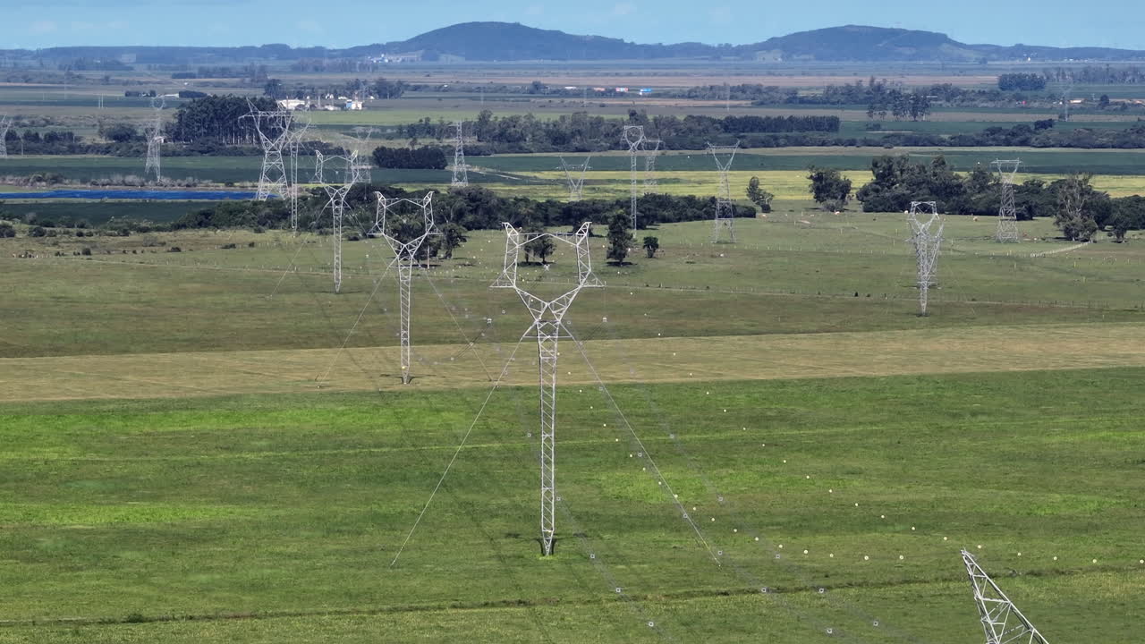 Torres de transmissão se estendem por campos abertos enquanto o drone sobe e inclina para baixo, enfatizando a infraestrutura de rede em grande escala e a distribuição de energia.