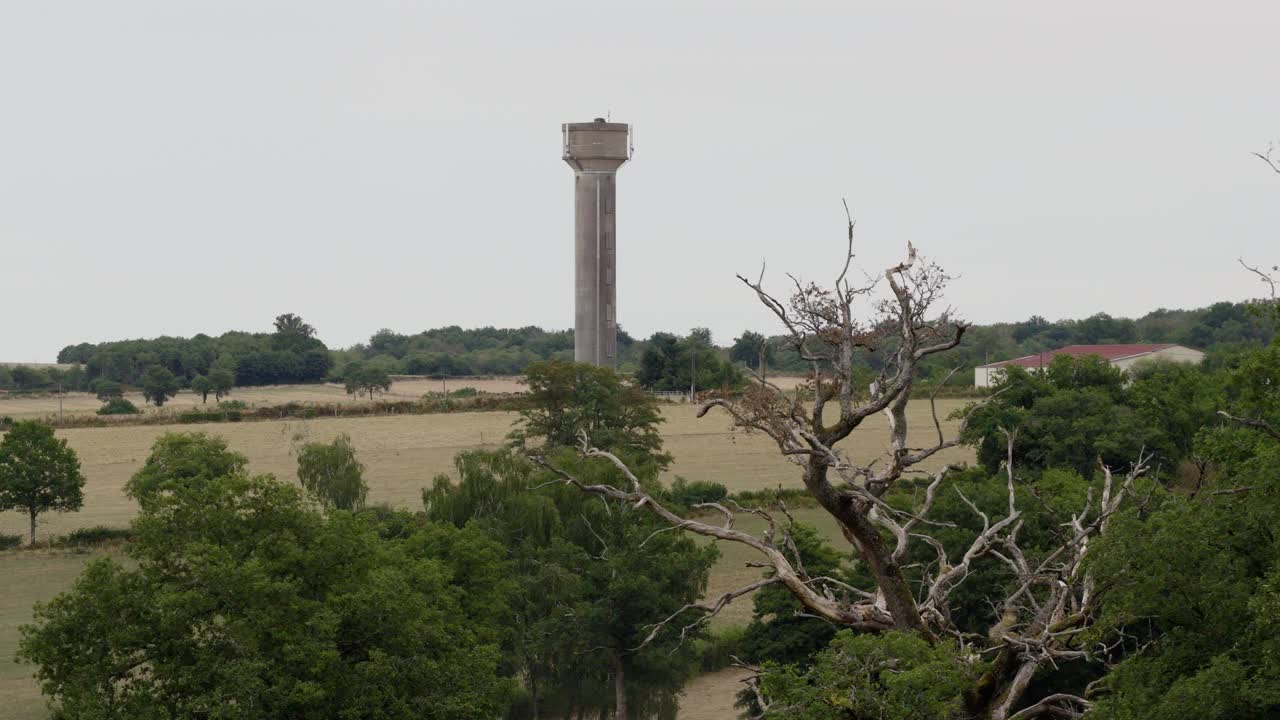 Ett högt vattentorn i betong står på landsbygden, omgivet av fält och träd, fångat i en bred flygbild