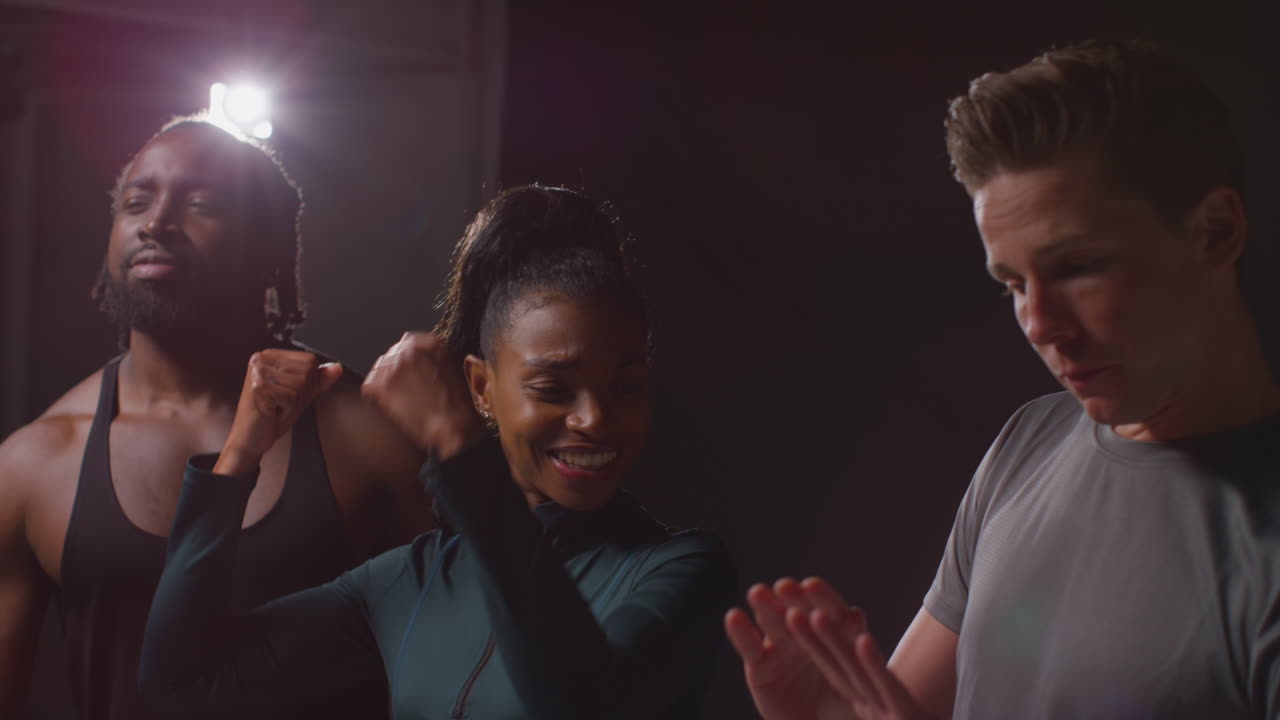 미소 짓는 남녀 운동선수들의 스튜디오 초상화, 피트니스 옷을 입고, 낮은 키 조명과 함께 체육관에서 즐겁게 포즈를 취하고 근육을 구부린다.