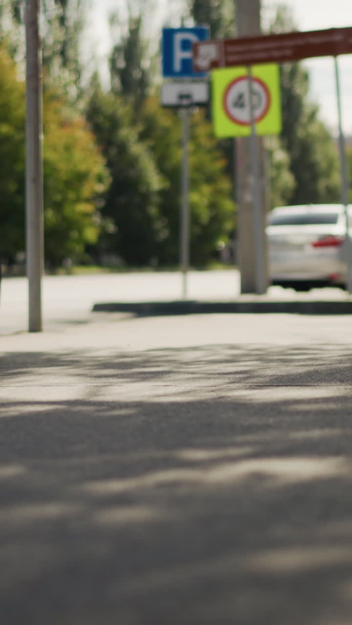 Ciclista circulando por una calle de la ciudad iluminada por el sol; un único ciclista con una chaqueta estampada pedalea junto a señales de aparcamiento y coches aparcados con sombras moteadas de árboles; un enfoque superficial en el asfalto enfatiza el movimiento y al viajero