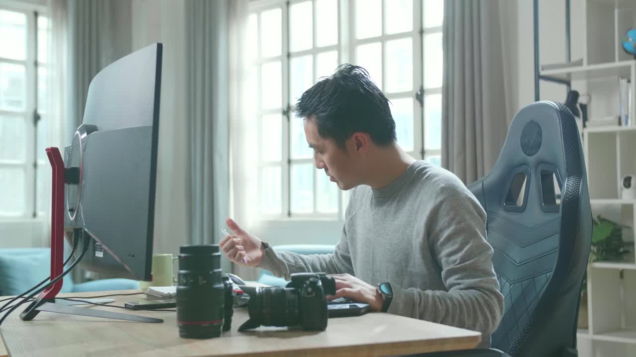 Asian Cameraman In Long Sleeved T-Shirt Looking At Desktop Computer Screen And Writing In Notebook On A Table While Working At Home.
