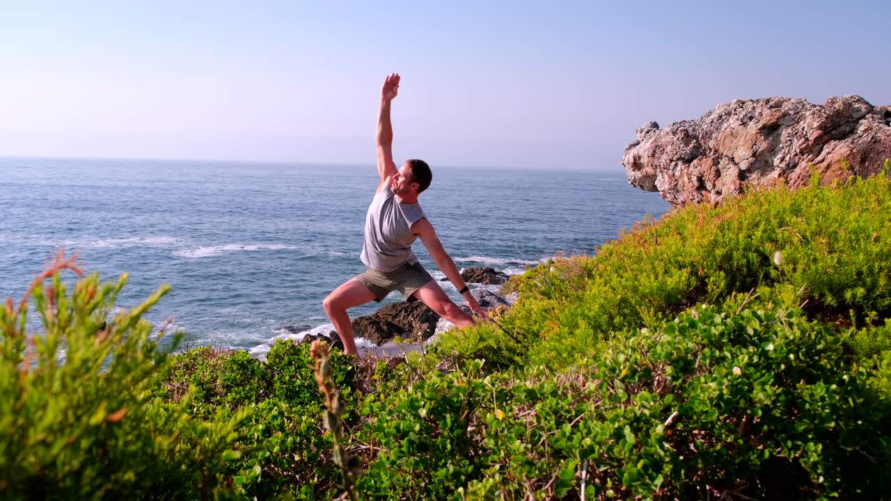 Fit man does yoga warrior poses on coastal cliff overlooking ocean at sunrise