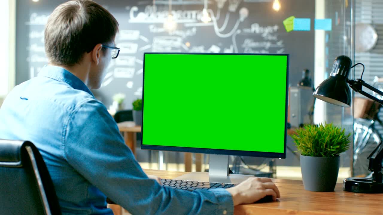 Young Man Works at His Desk on the Personal Computer with Mock-up Green Screen. In the Background His Colleague Works in the Creative Office.