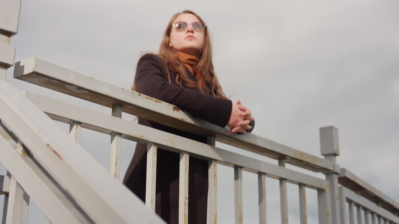 Girl in glasses and trench coat calmly observes surroundings while leaning on metal handrail under calm weather, showing tranquility thoughtful posture, and quiet awareness in natural outdoor light