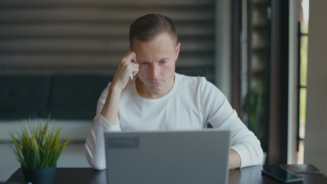 un joven con una pluma en la mano se sienta en la habitación en el escritorio con una computadora portátil y mira pensativo a la pantalla. la mano derecha frota el templo