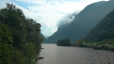 a tranquil river flows between lush green banks and majestic mountains, partially covered by clouds. the calm waters reflect the natural beauty of the surroundings