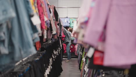 woman chooses children clothes in store. lady in denim suit seeks stylish outfit for kids in large fashion shop. customer stands in aisle between racks