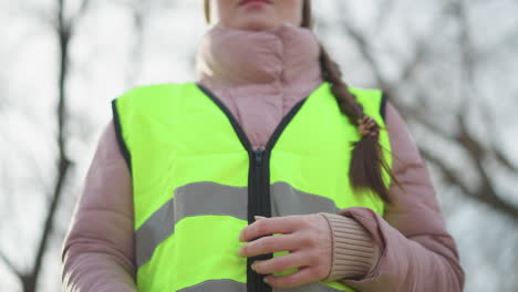 close-up of woman in pink puffy jacket zipping up bright reflective safety vest outdoors on cool day, preparing for work or volunteering, long braid visible over shoulder, blurred trees in background