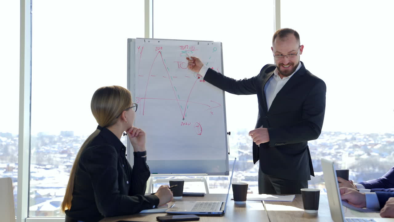 Smiling businessman standing near board and talking. Man leader in suit explaining company development strategy to colleagues showing on painted graphics.