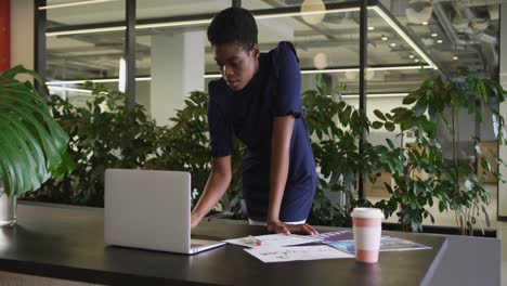 African-american-businesswoman-standing-using-laptop-going-through-paperwork-in-modern-office