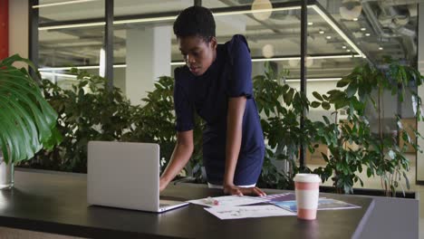 african american businesswoman standing using laptop going through paperwork in modern office