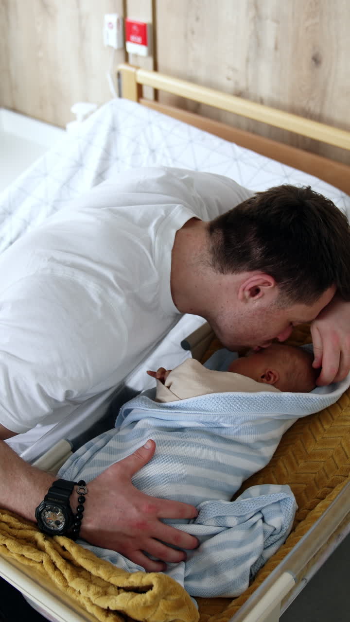 Loving dad sits at the crib with his baby. Happy father leans to kiss his newborn child on the head. Vertical video