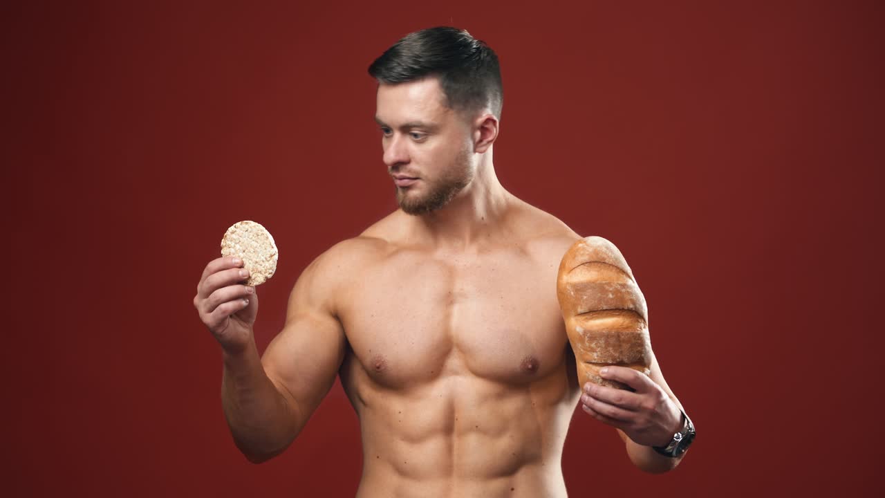Healthy food choices. Naked sportsman choosing between healthy and unhealthy bread for his muscular body. Portrait of handsome bodybuilder with bread in studio.