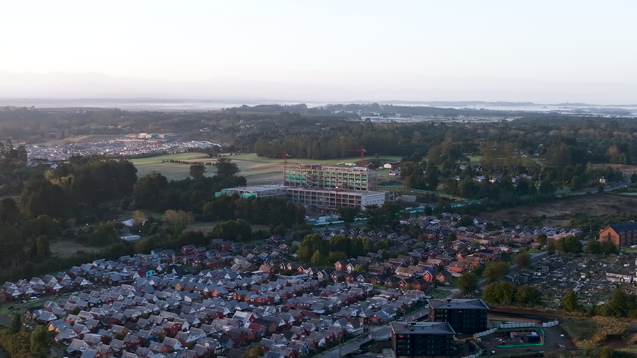 Luftaufnahmen einer Drohne bei Sonnenaufgang, die die Baustelle des zukünftigen Krankenhauses in Puerto Varas, Chile, mit Kränen und Morgenlicht zeigen.