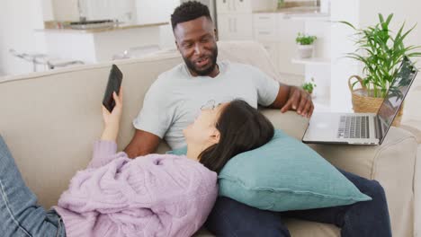 diverse couple lying on couch and using laptop in living room