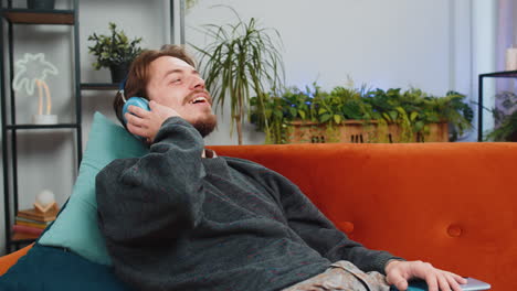 hombre feliz con auriculares inalámbricos relajándose escuchando su música rock n roll favorita acostado en el sofá de casa