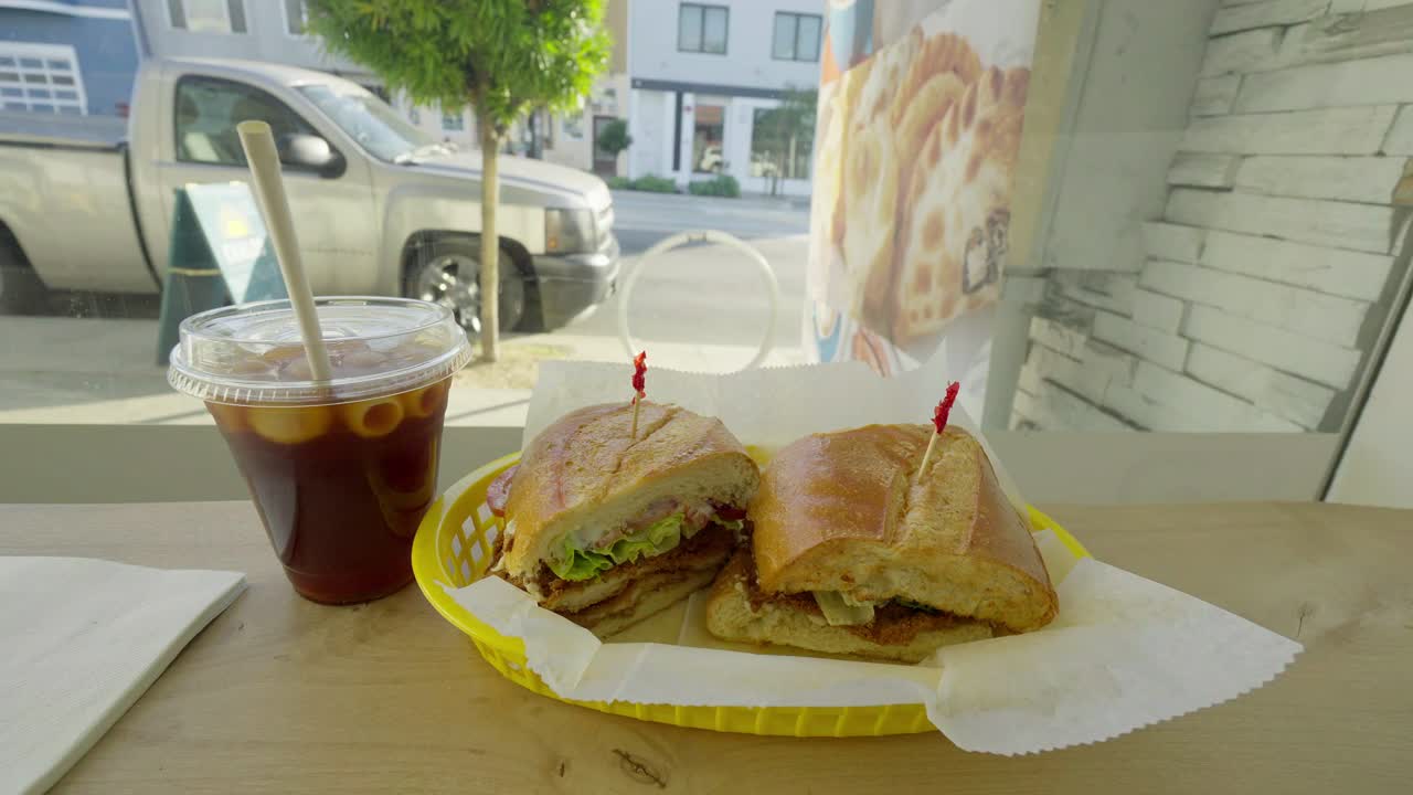 A chicken sandwich rests beside an iced coffee on a sunlit table, window light casting soft shadows that create a relaxed café mood perfect for casual dining visuals