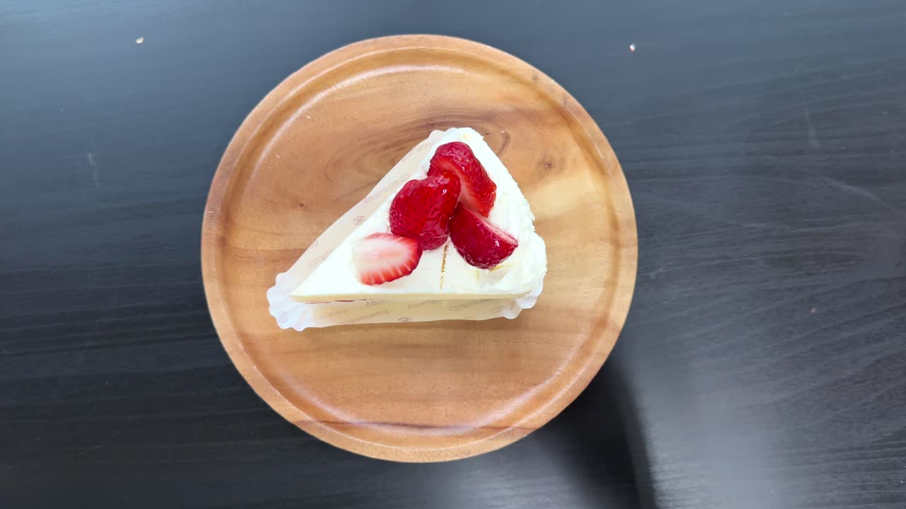 Hand places birthday candle on strawberry shortcake slice, wooden plate, bright overhead lighting, static shot
