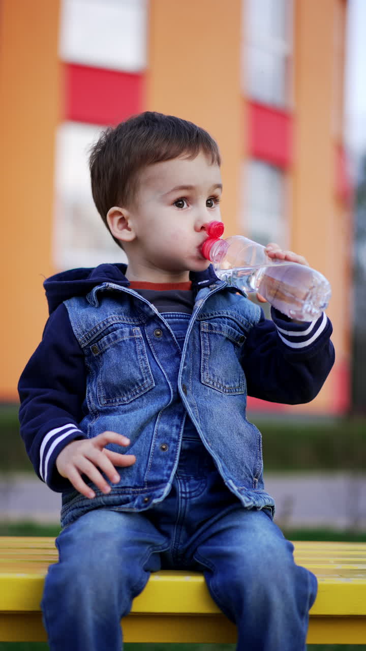 Caucasian toddler in jeans romper and jacket sits on the bench. Kid drinks water looking ahead thoughtfully. Vertical video.