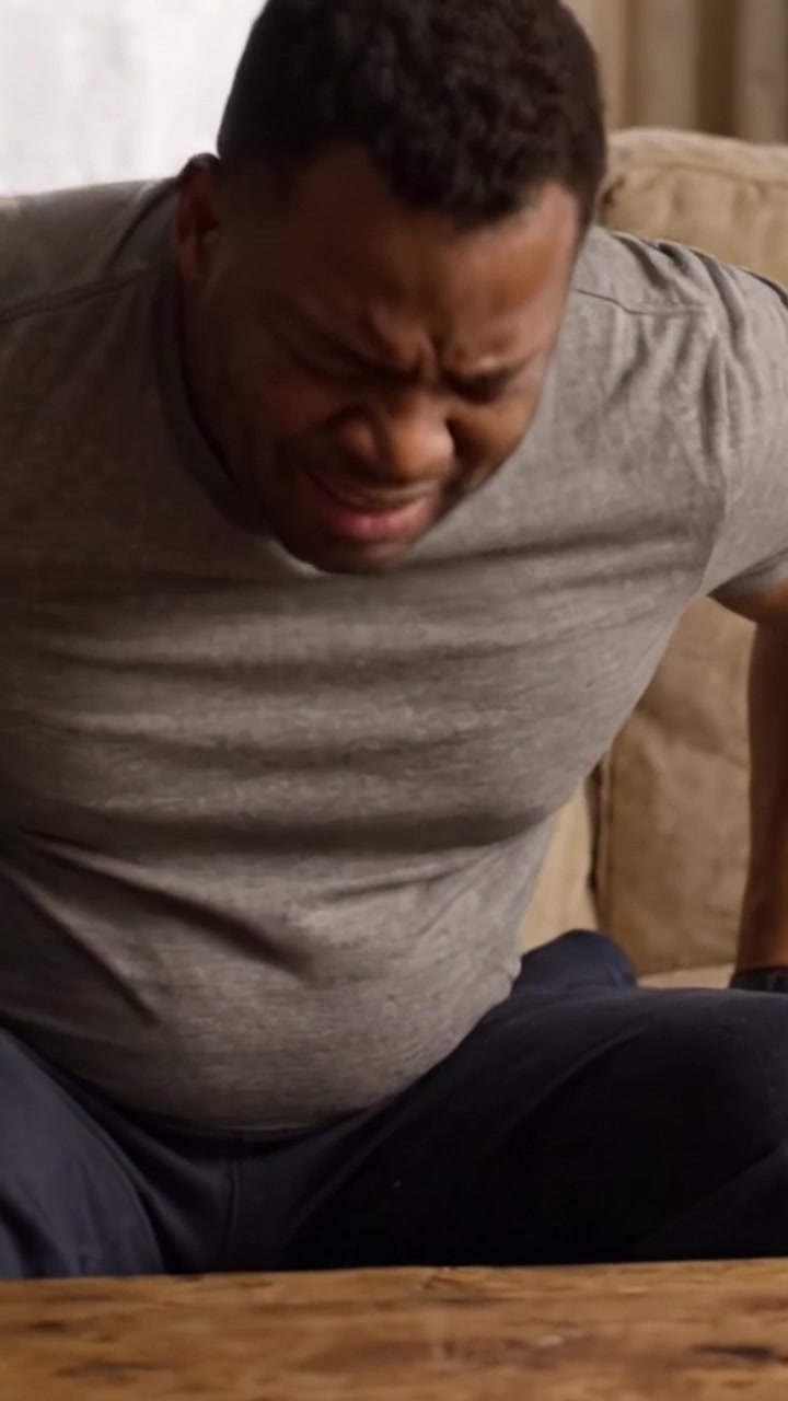A Man Struggles with Discomfort and Pain While Sitting on a Couch, Reflecting Physical Distress and Emotional Turmoil in a Domestic Setting