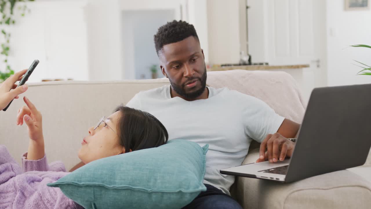 Diverse couple lying on couch and using laptop in living room