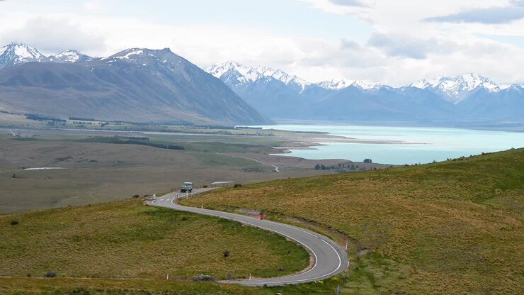 Camion agricolo che percorre una strada tortuosa con una splendida catena montuosa alpina sullo sfondo e il lago Tekapo turchese.