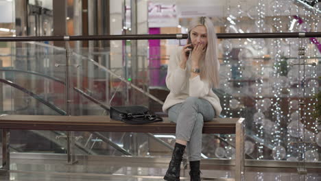 young woman seated on mall bench, leaning forward with phone held to ear, face showing exhaustion, wearing sweater, jeans and boots, festive lights and glass railing in background, purse beside her
