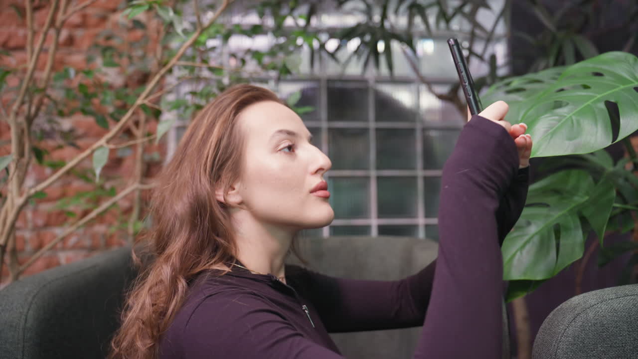Side view of blond woman with soft smile focusing on phone while snapping photo near indoor plants, seated in modern green booth with natural lighting and lush leafy background