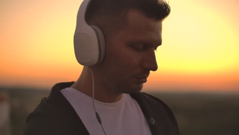 smiling and laughing handsome young man listening music from his smartphone in wireless headphones. in slow motion a man stands on the roof at sunset and looks at the beautiful view from the height of the city