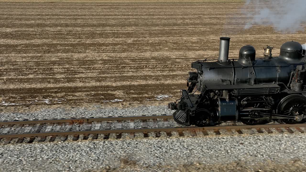 A vintage steam locomotive chugs along railway tracks surrounded by golden fields, puffing smoke