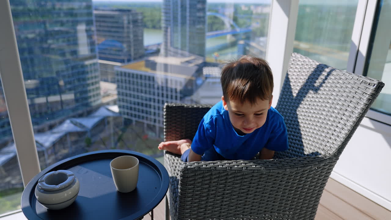 Child leaning on balcony chair. A boy in a blue shirt leans on a wicker chair on a high balcony overlooking tall buildings and a river