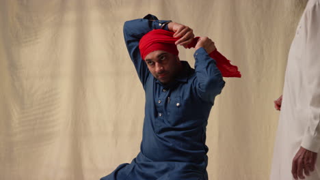 Studio-Shot-Of-Senior-And-Younger-Sikh-Men-Tying-Fabric-For-Turban-Against-Plain-Background