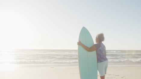 happy senior african american woman holding surfboard at beach, in slow motion, with copy space