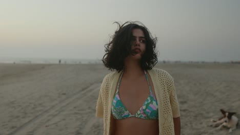 Young-woman-in-bikini-and-cover-up-standing-on-a-beach-at-dusk,-with-a-dog-in-the-background