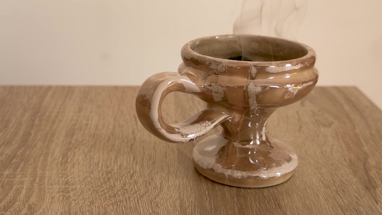 Un quemador de incienso de cerámica esmaltada marrón libera finas espirales de humo mientras descansa sobre una mesa de madera, enmarcado contra una pared interior beige lisa en una toma estática cercana.