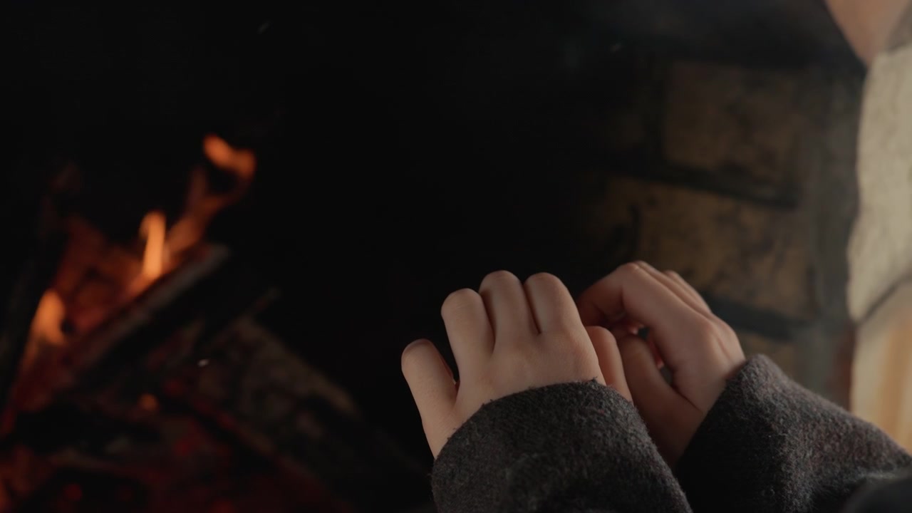 Mani a coppa si muovono vicino a braci ardenti e piccole fiamme all'interno di un focolare rustico in pietra, la morbida luce del fuoco proietta calde ombre arancioni su maniche, dita e palmi in un tranquillo comfort invernale.