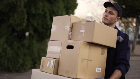 handsome delivery man in uniform, cap and gloves carrying many heavy cardboard boxes parcels outdoor