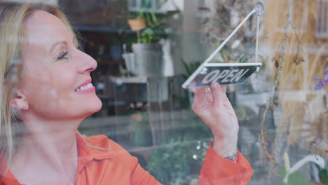 sonriente mujer madura propietaria de una pequeña empresa girando alrededor de un letrero abierto en la ventana de la tienda