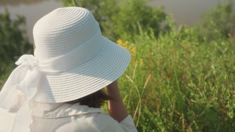 close up rear view of woman in long white sleeve bending toward wild yellow flowers while photographing them, surrounded by green bush and blurred river in background