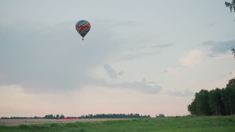 open grass field with distant hot air balloon drifting overhead during serene sunset sky while ground crew prepares next launch against trees and pastel clouds