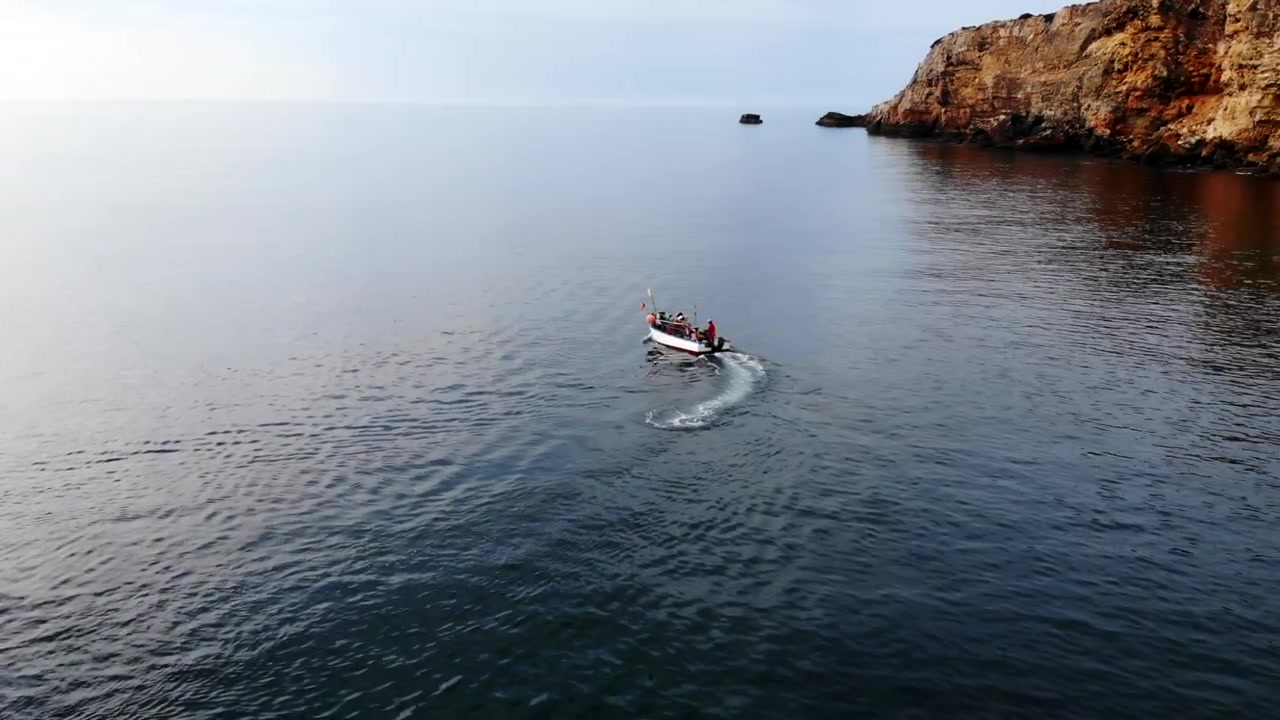 Barca da pesca vicino a scogliere, equipaggio che naviga su acqua calma e riflettente lasciando una scia stretta, ampio orizzonte. Media