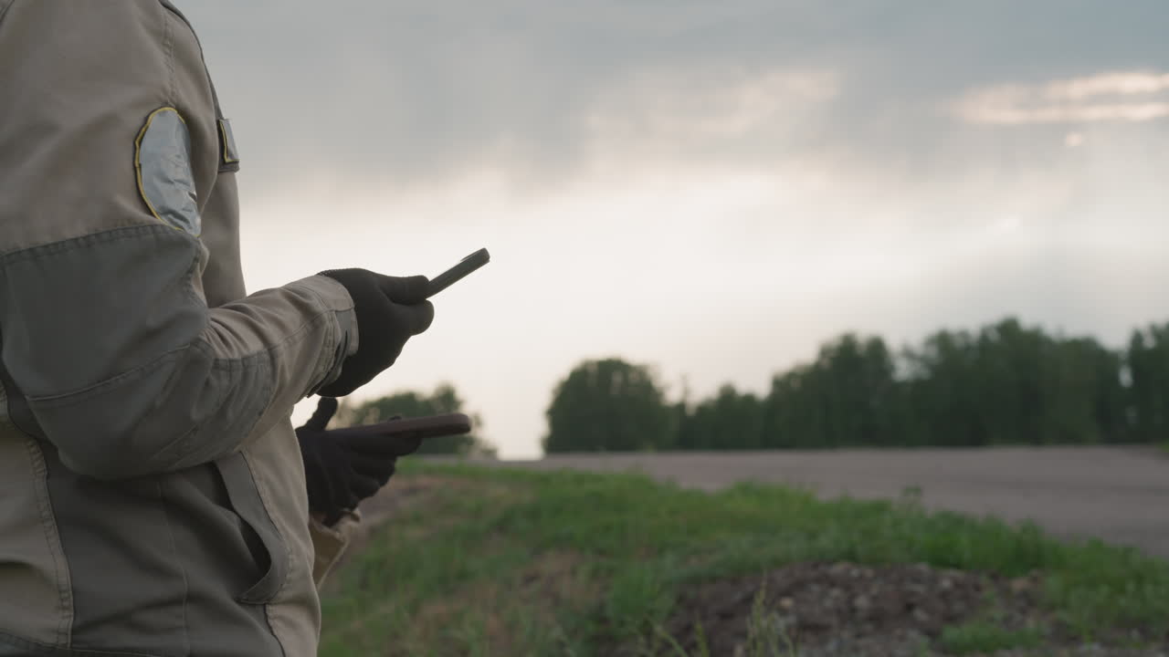 close up of two field workers holding handheld devices beside country road under moody sky, wearing outdoor gear and gloves, hinting collaboration, weather monitoring