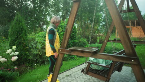 woman in yellow and green jacket with yellow boots walks toward mint-colored bag resting on swing bench, background features lush greenery, garden, trees, and a plastic water drum