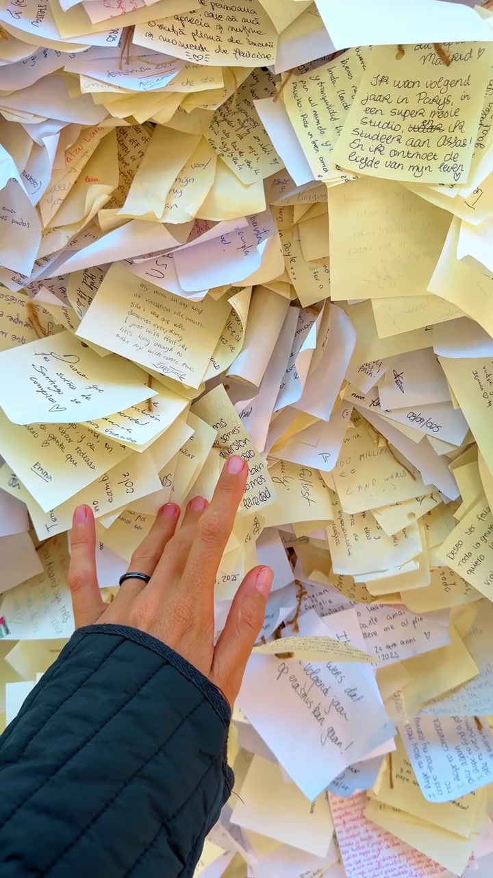 Madrid, Spain, 8 August 2025: Hand of person touching wall with many paper notes. Close up of human hand over collection of handwritten messages and wishes pinned on public display board.