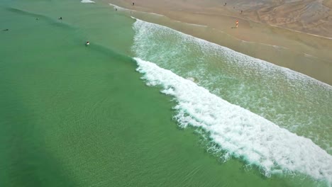 Vista-De-Arriba-Hacia-Abajo-De-La-Playa-Fistral-Con-Movimiento-Lento-De-Olas-Y-Surfistas-A-Lo-Largo-De-La-Playa,-Aguas-Turquesas-En-Cornualles,-Reino-Unido