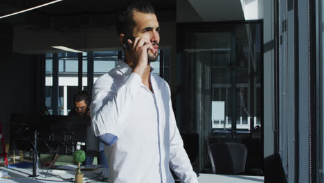 hombre de negocios de raza mixta de pie en una oficina soleada hablando en su teléfono inteligente y mirando por la ventana