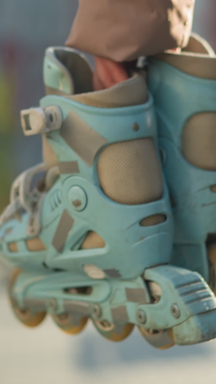 Close-up of a person holding a pair of rollerblades, wearing black trousers, walking through a park with a blurred background of trees and people, capturing a casual and leisurely moment