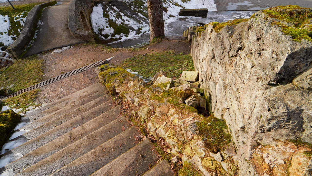 Worn stone stairs beside a damaged moss-covered wall with loose masonry, showing historic decay, unsafe edges, and spring sunlight in an old outdoor setting.
