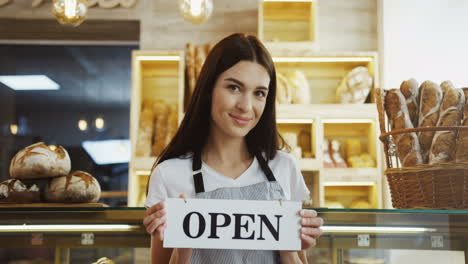 Female-Smiled-Bakery-Vendor-Putting-And-Turning-Signboard-Open-On-The-Glass-Door-Of-The-Bakery-Shop